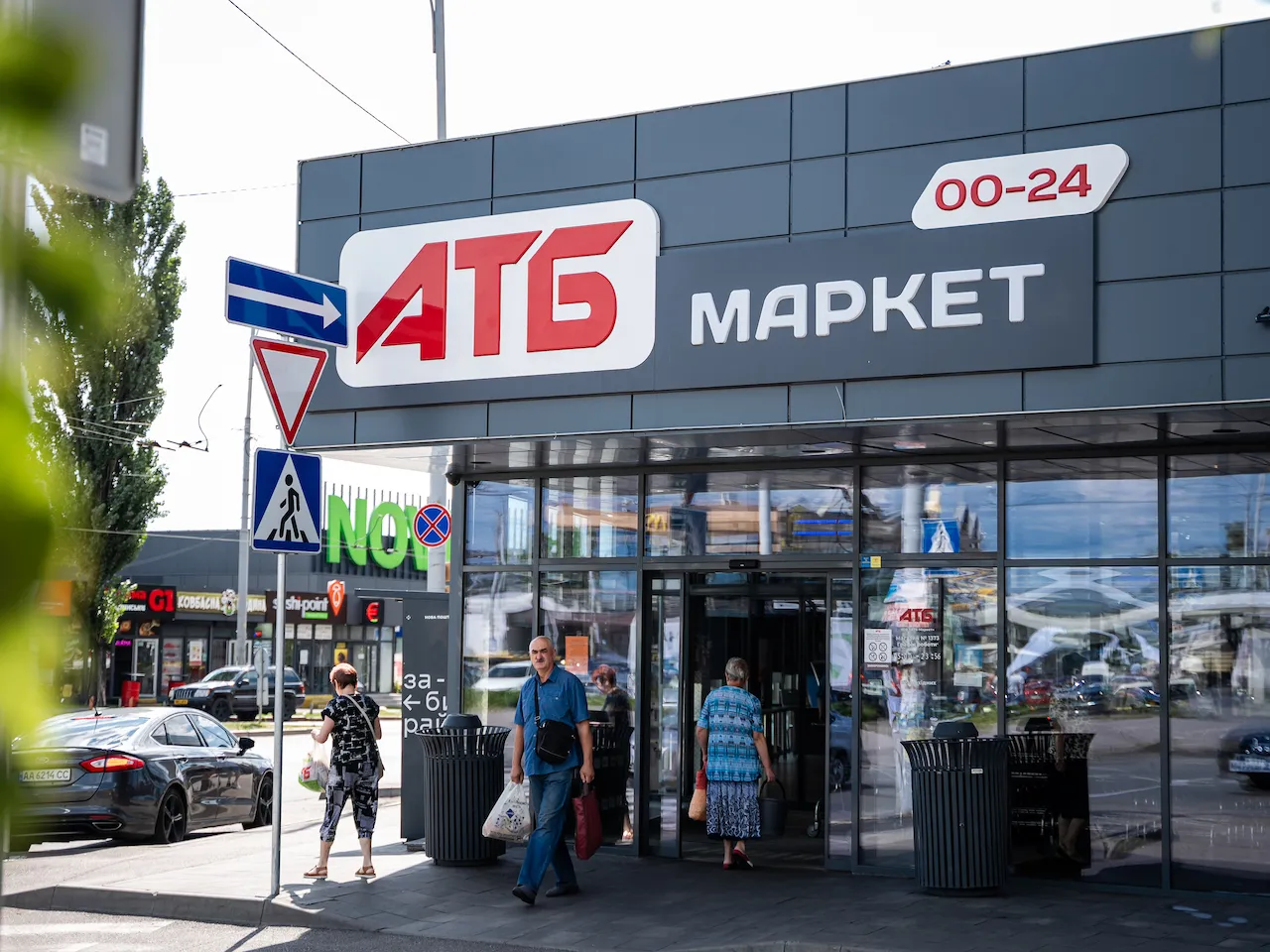 Вхід до маркету АТБ з людьми, які виходять і заходять, поруч з вуличними знаками і авто.