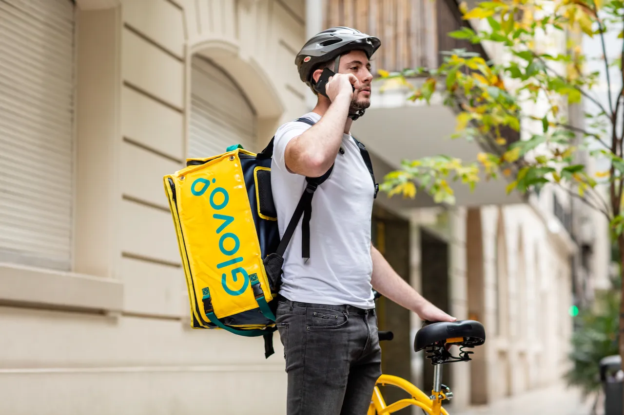 Кур'єр Glovo з жовтим рюкзаком стоїть біля велосипеда і розмовляє телефоном.