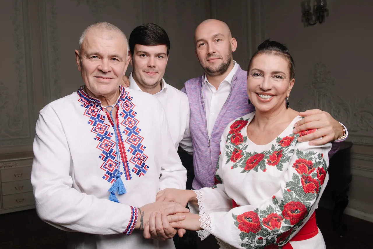 Четверо людей у вишиванках і сорочках усміхаються, тримаючись за руки в кімнаті з класичним декором.