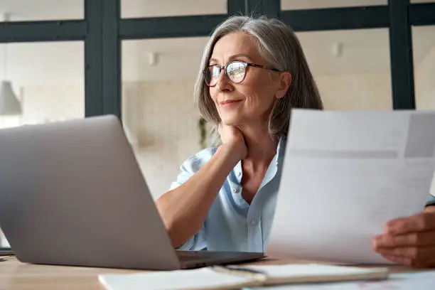 Mature woman with glasses sitting at a desk, looking thoughtfully at her laptop while holding a document.