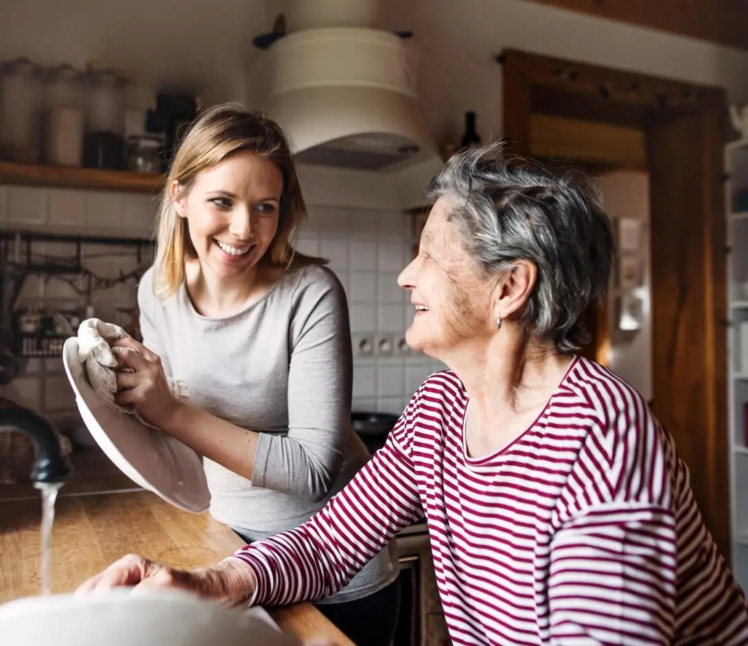 Young woman drying a plate and smiling at an elderly woman in a kitchen.