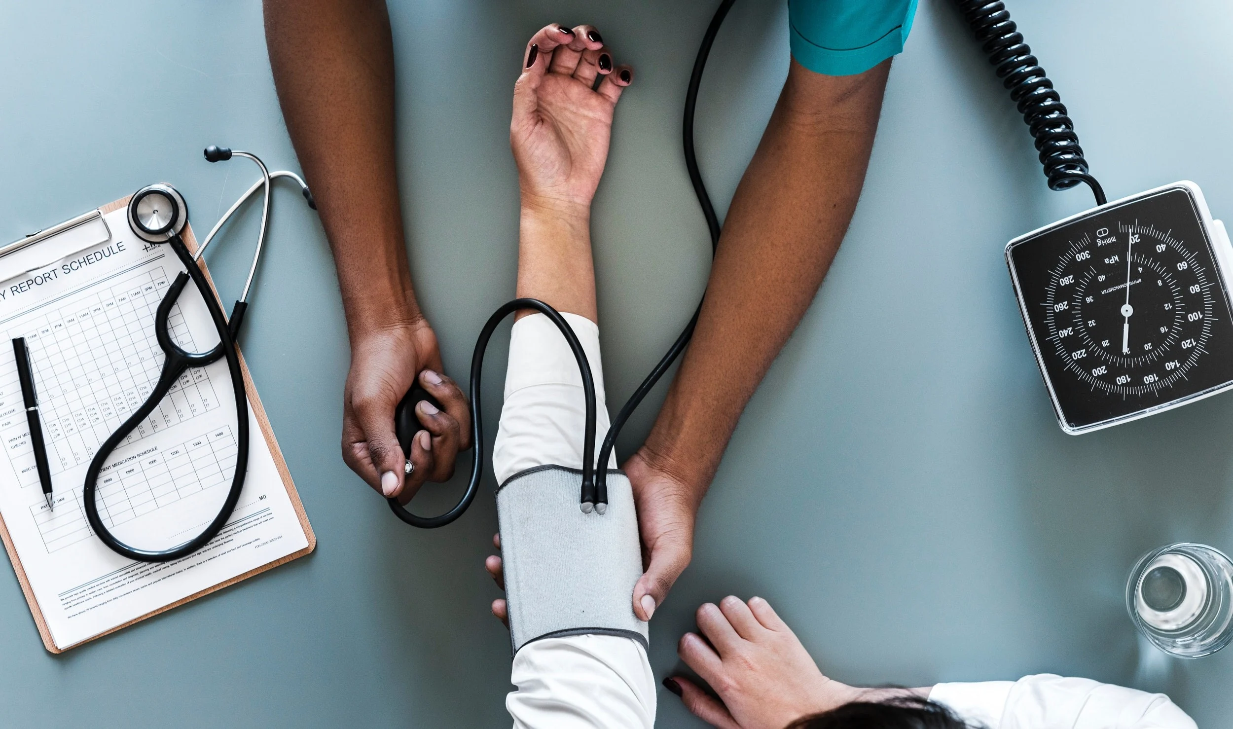 Healthcare professional measuring a patient's blood pressure with a sphygmomanometer and stethoscope on a gray table.