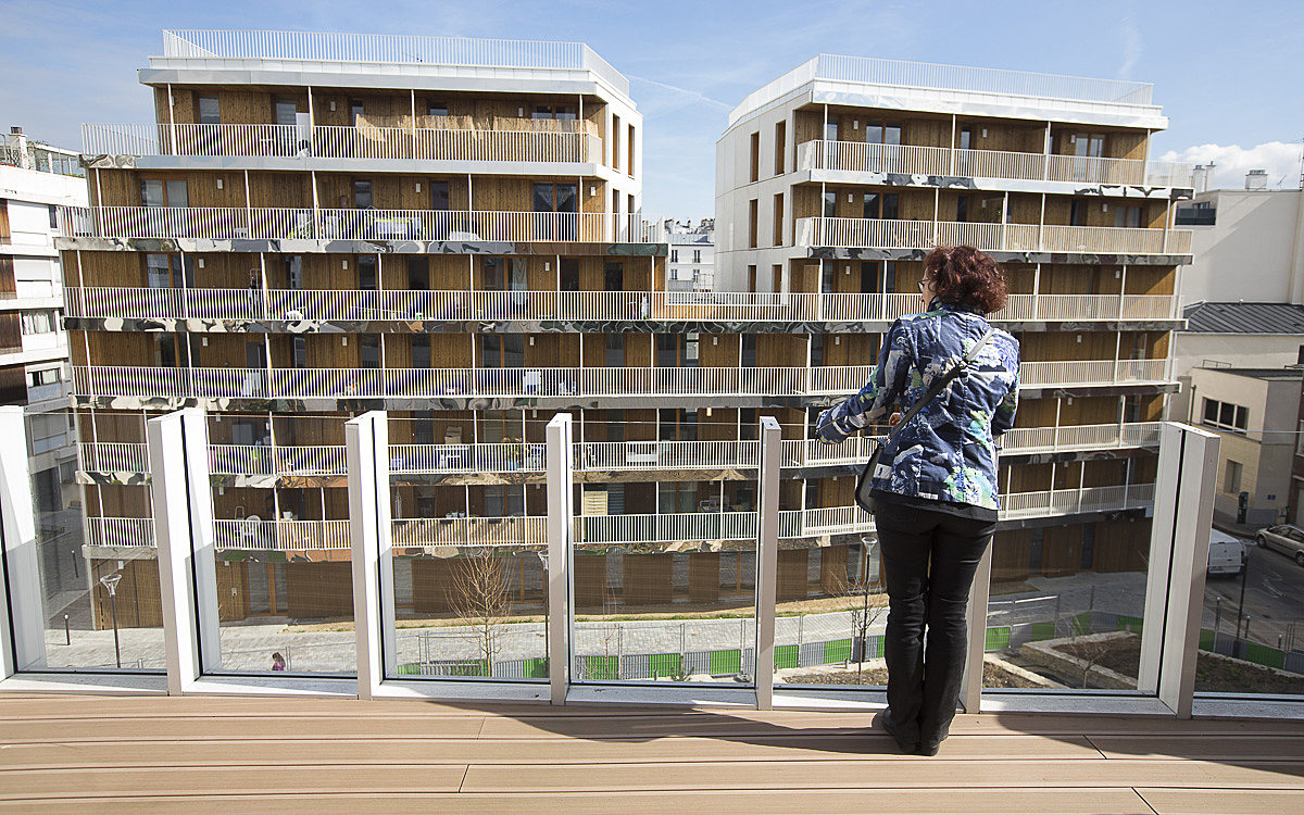 Person with curly hair and blue patterned jacket standing on a wooden balcony, overlooking modern apartment buildings with glass railings and wooden panel balconies.