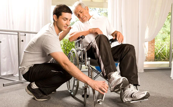 Young man assisting an elderly man in a wheelchair by adjusting his footrest indoors.