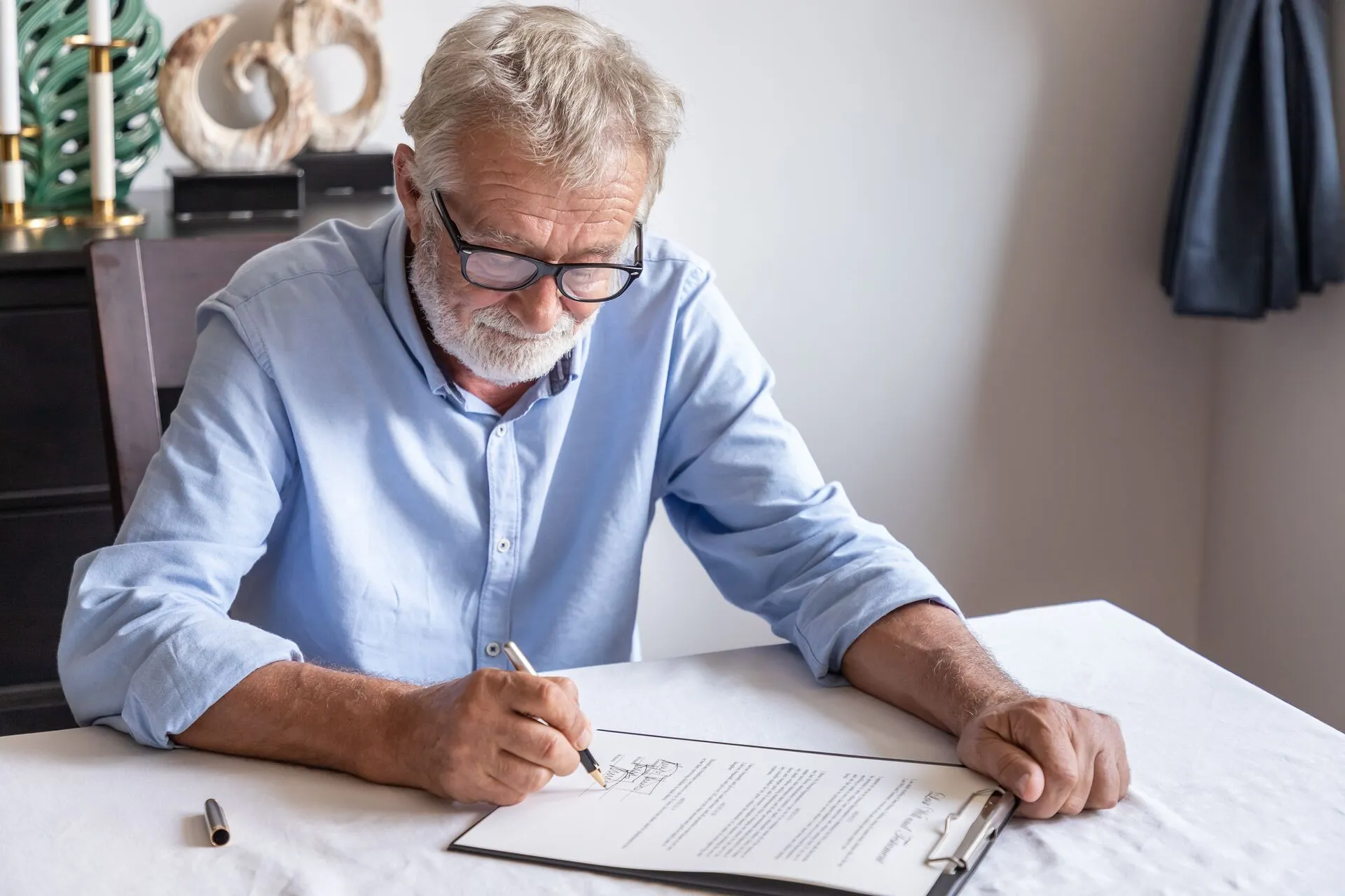 Elderly man in glasses and blue shirt signing a document on a clipboard at a table.