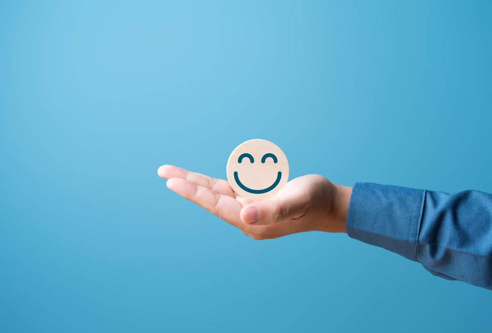 Hand die een houten schijf met een glimlachend gezicht vasthoudt tegen een blauwe achtergrond.