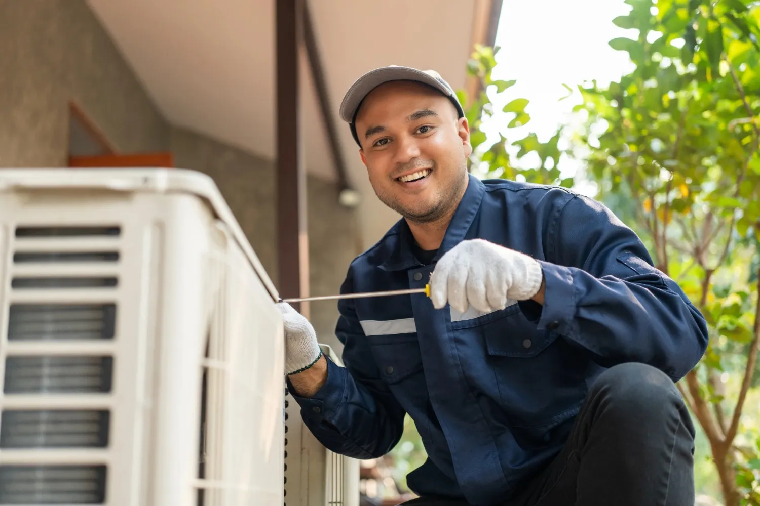 A smiling technician wearing cap is repairing an outdoor AC unit using tools 