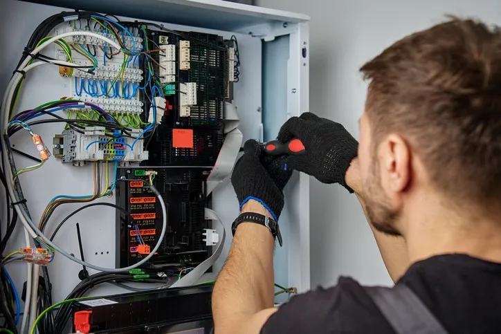 A technician wearing black gloves uses a screwdriver to work on the complex electrical wiring and circuit boards inside an open heat pump control panel. Blue, yellow, and green wires are visible throughout the internal system.