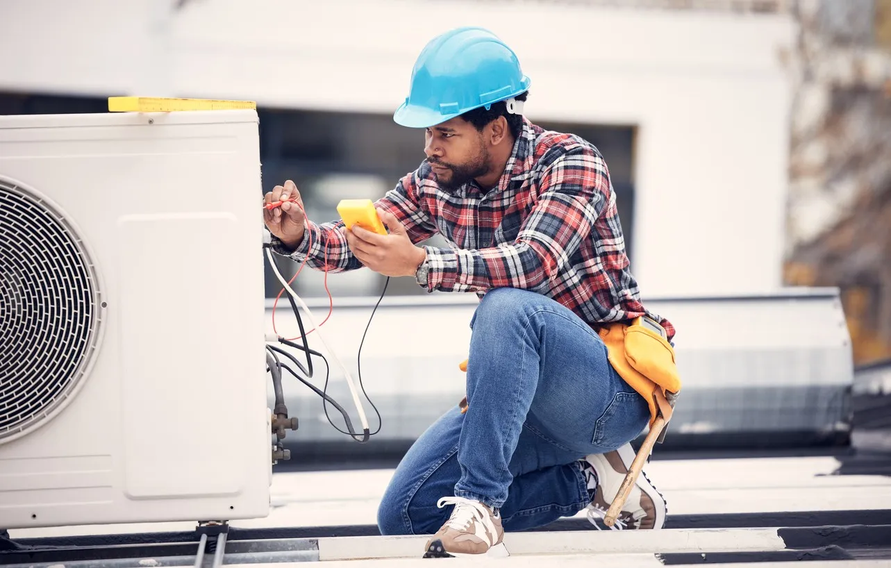 A technician wearing a blue hard hat, plaid shirt, and jeans kneels on a flat roof to test a white outdoor heat pump unit. He is using a yellow multimeter to check the electrical connections.