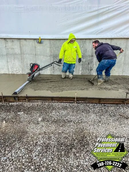 Two concrete contractors are actively working on a freshly poured concrete slab, one using a power screed and the other a float, with a business logo in the bottom right corner.