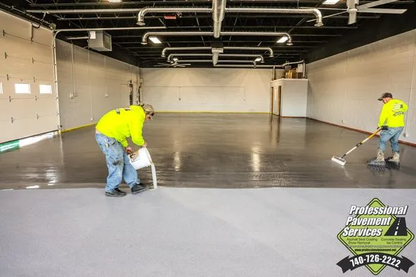 Two workers apply an epoxy coating to a large gray floor in a spacious garage or warehouse, with a company logo in the bottom right corner. Analysis