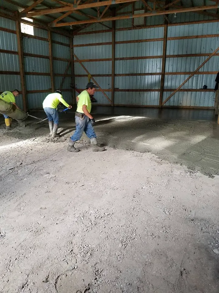 Three workers in a barn-like structure are installing a concrete floor, with one worker using a chute to pour concrete. Show thinking