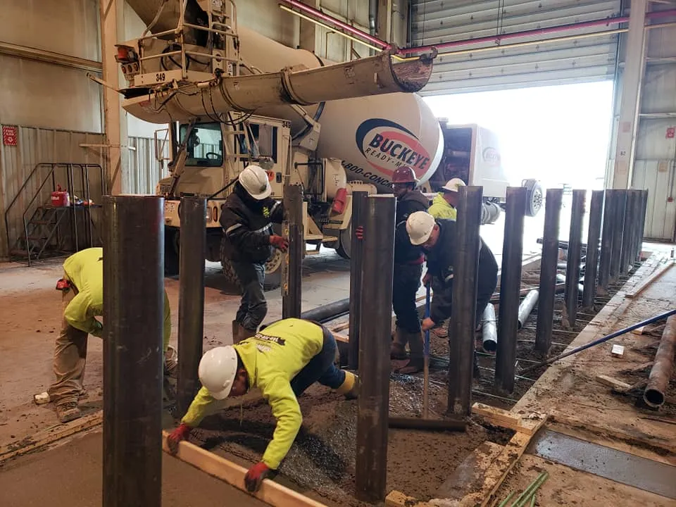 A team of concrete contractors are installing bollards, pouring concrete around them with the help of a ready-mix truck in the background.