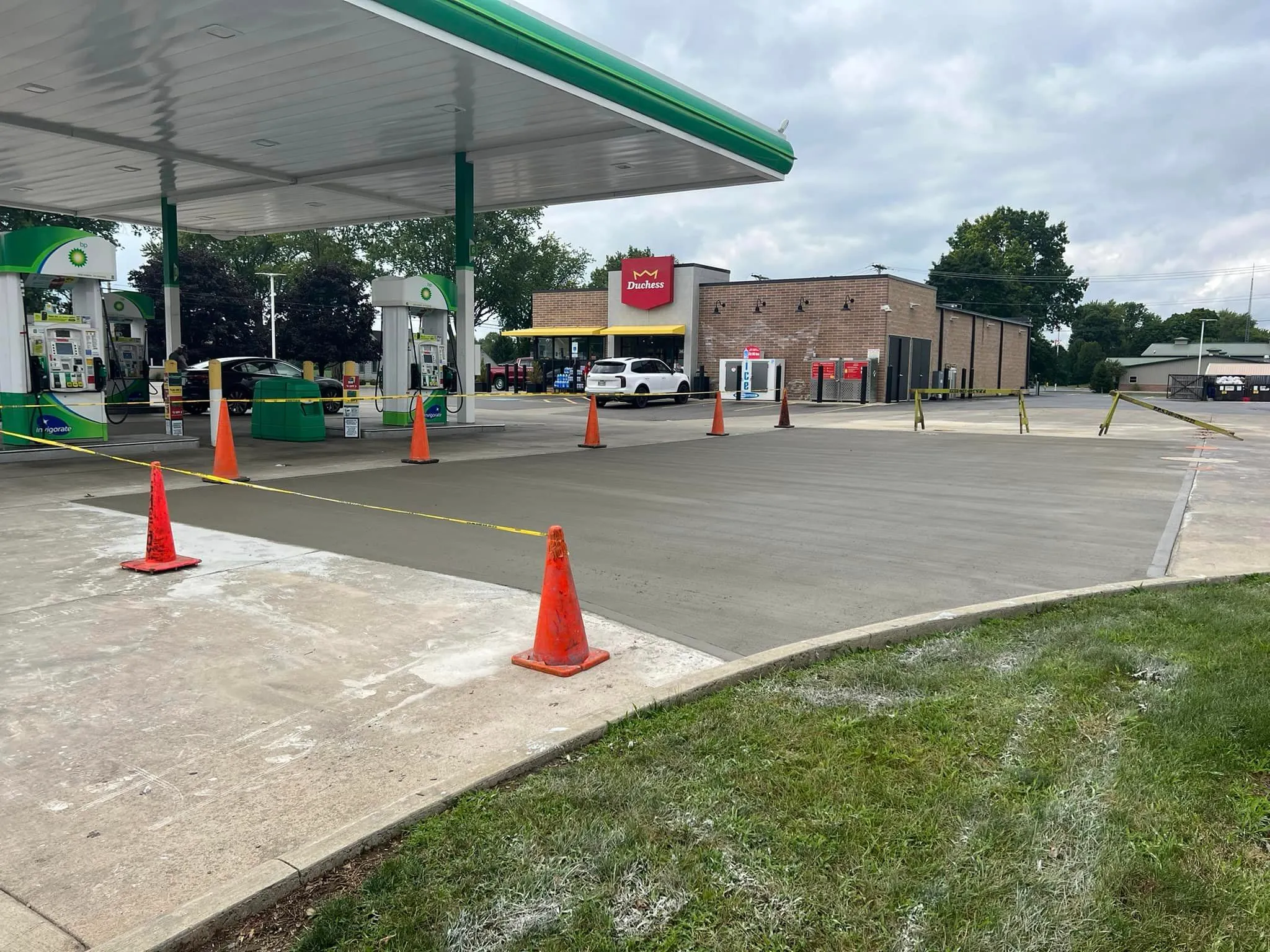 A freshly poured concrete pad, marked with orange cones and yellow caution tape, sits under the canopy of a gas station on a cloudy day.