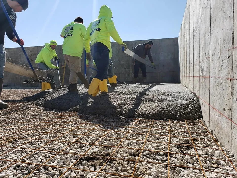 A group of concrete contractors are pouring and leveling concrete over a rebar grid and gravel base next to a concrete wall on a sunny day.