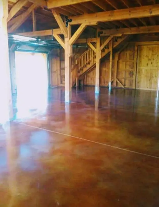 A rustic interior space features a highly polished, richly stained concrete floor with warm brown and amber tones, reflecting the wooden beams and staircase within.