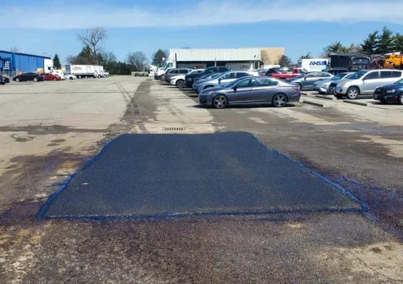 A large, square-shaped patch of freshly laid asphalt is shown in the middle of a worn, commercial parking lot.     
