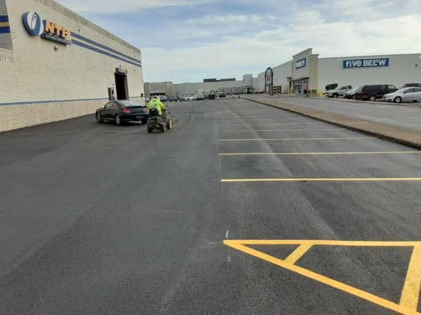A worker is driving a line-striping machine to paint a parking lot with yellow lines outside of a commercial plaza with stores like NTB and Five Below.