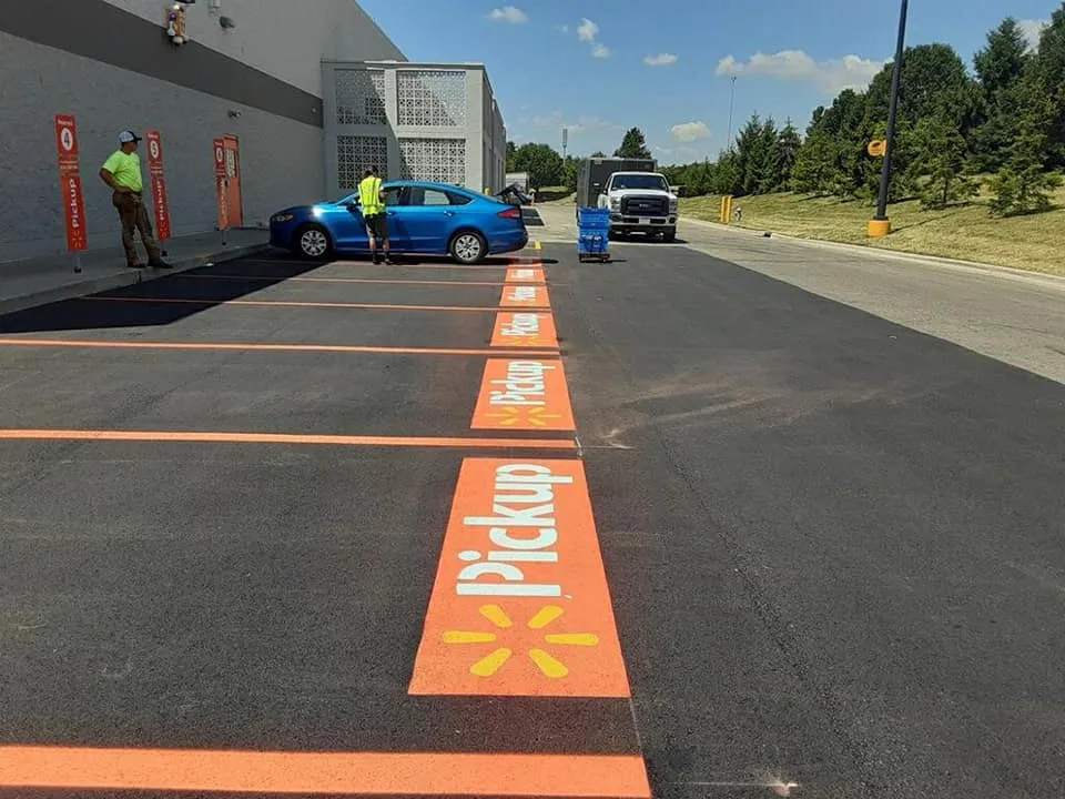 Freshly painted orange "Pickup" parking spaces with a Walmart logo are seen in a parking lot, with two workers and a blue car nearby.