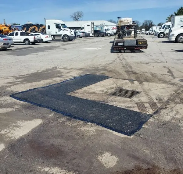 A large, square-shaped patch of freshly laid asphalt is shown in the middle of a worn, commercial parking lot.        