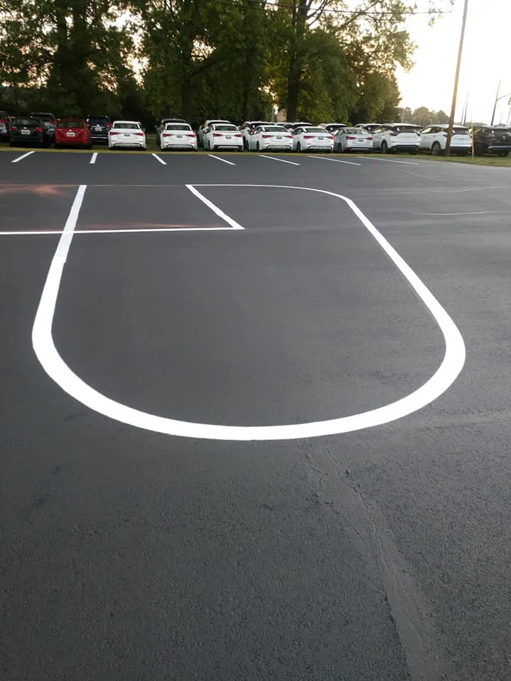 A newly paved and sealed parking lot features a large, crisp white U-shaped line in the foreground, with cars parked in the background.