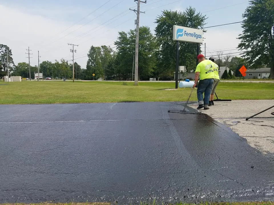 Two workers are applying a black sealcoating to a commercial parking lot with a "Ferrellgas" sign visible in the background.