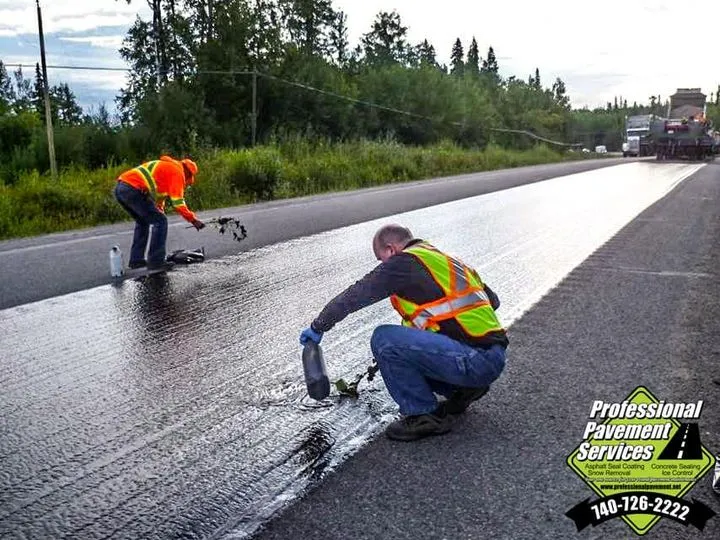 Two workers in neon vests are applying sealcoating to an asphalt road, with one pouring from a bottle and the other raking the liquid.
