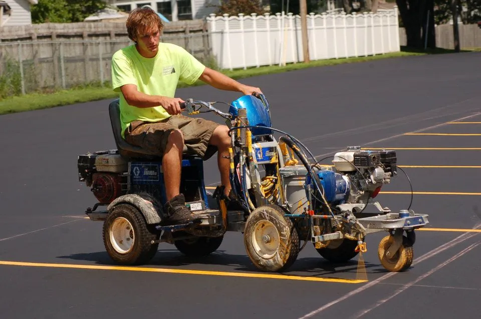 A worker is shown on a small ride-on machine painting yellow lines on a newly paved black asphalt parking lot.