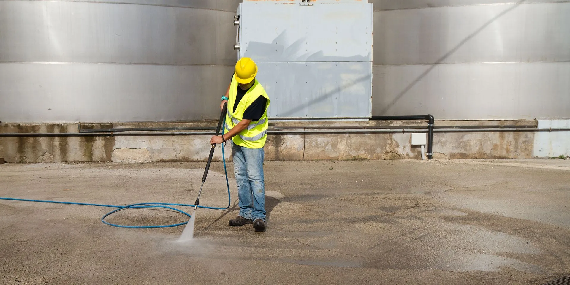 A construction worker wearing a hard hat and safety vest uses a power washer to clean a concrete slab in front of a large metal building.