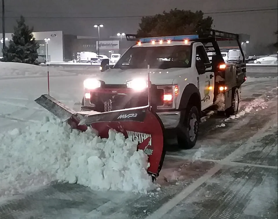 A professional snow removal truck with a large plow pushes a pile of snow to the side of a parking lot at night.