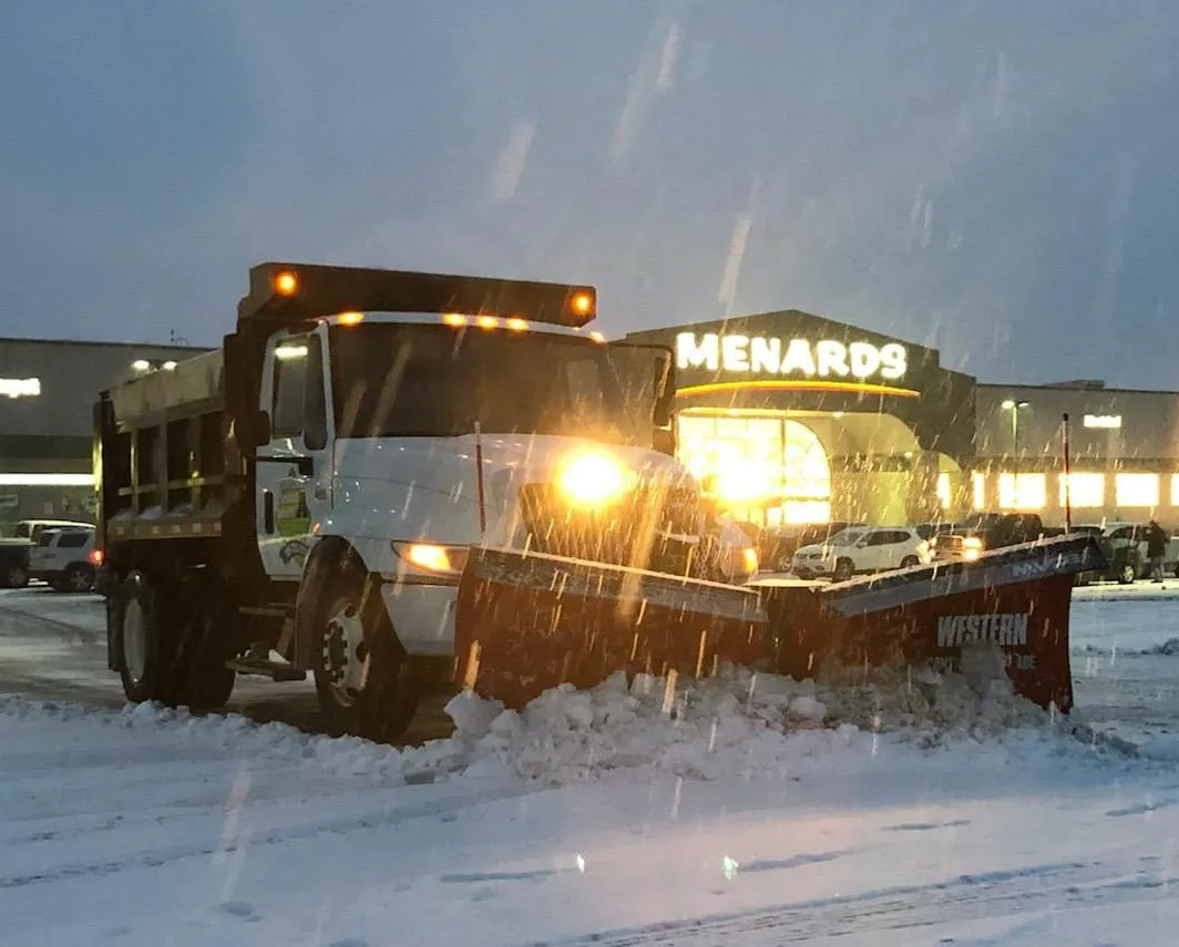 A close-up view of a snowplow blade pushing a large wave of snow at night.