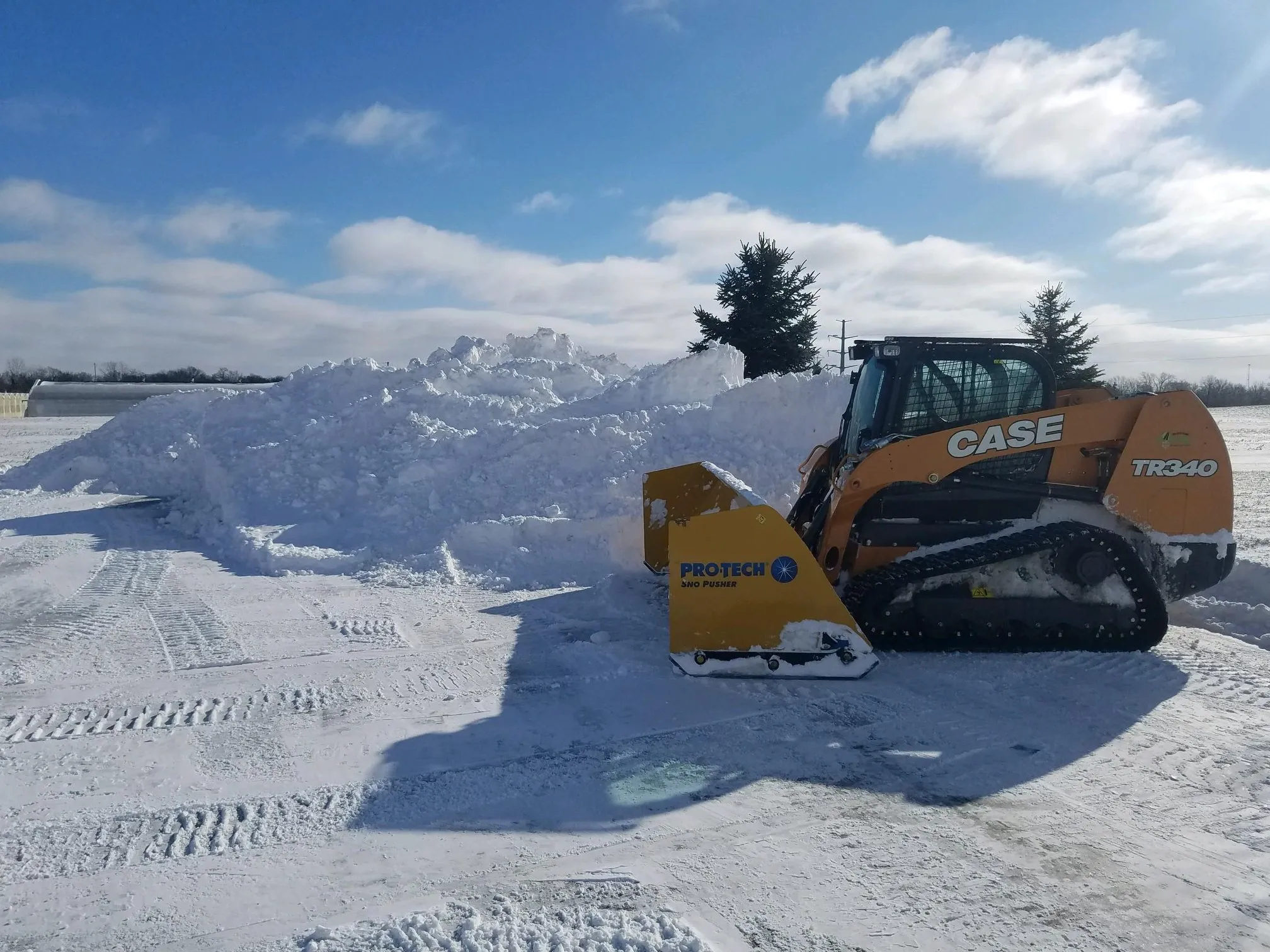 A tracked CASE TR340 loader with a yellow "PROTECH" snowplow attachment pushes a large pile of snow on a bright, sunny day.