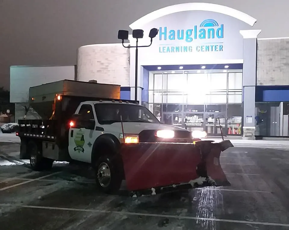 A large white snow removal truck with a red plow and a salt spreader on the back is parked in front of a building with a sign that says "Haugland Learning Center" at night.