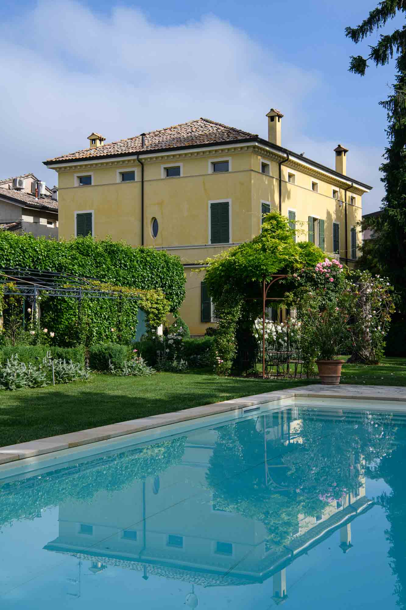 Tranquil swimming pool reflecting the historic yellow villa, surrounded by a blooming Italian garden and curated hedges.