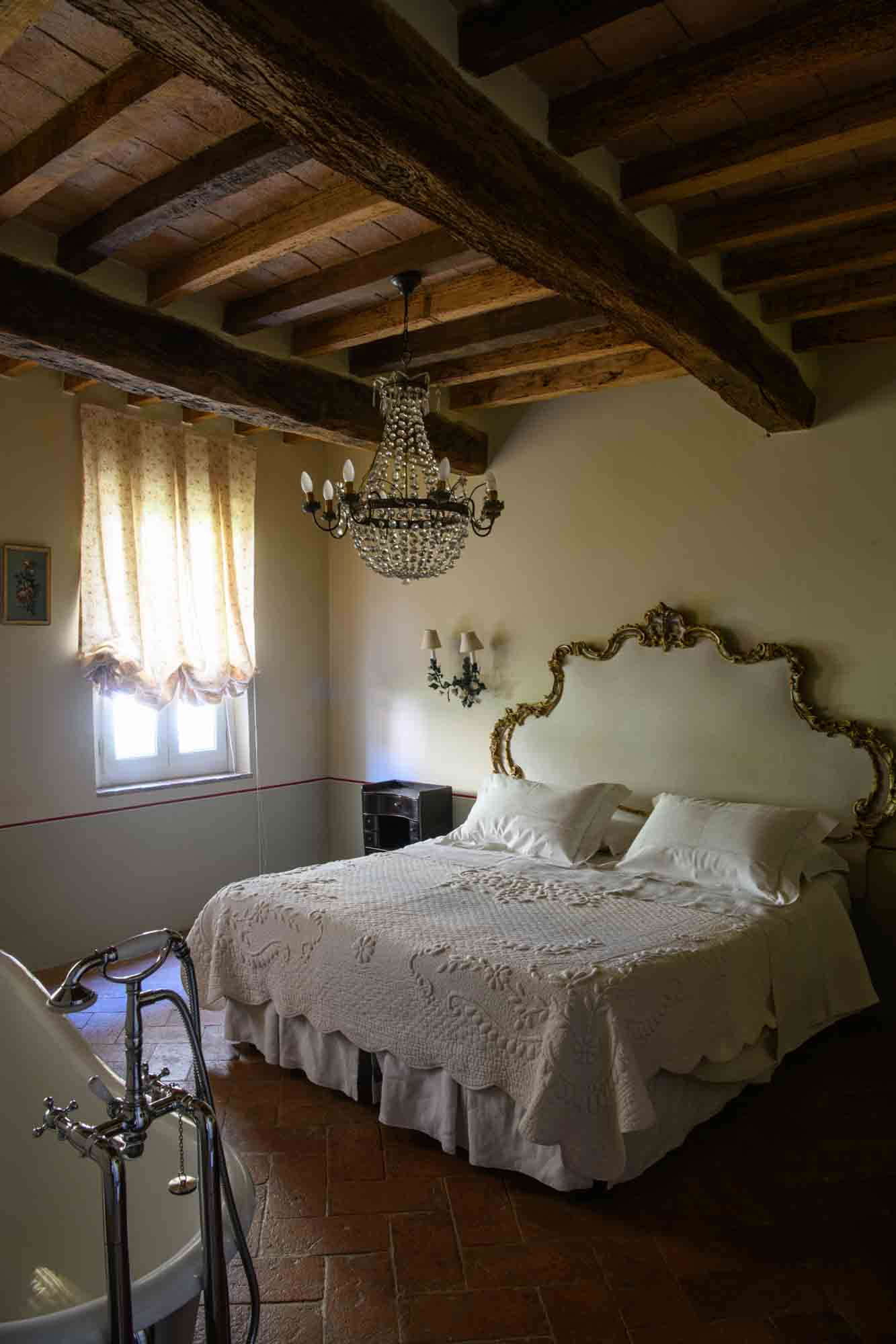 legant country-style bedroom in Italy featuring antique gold headboard, freestanding bathtub, vintage chandelier, and exposed wooden beams – a refined space for exclusive stays and authentic local experiences.
