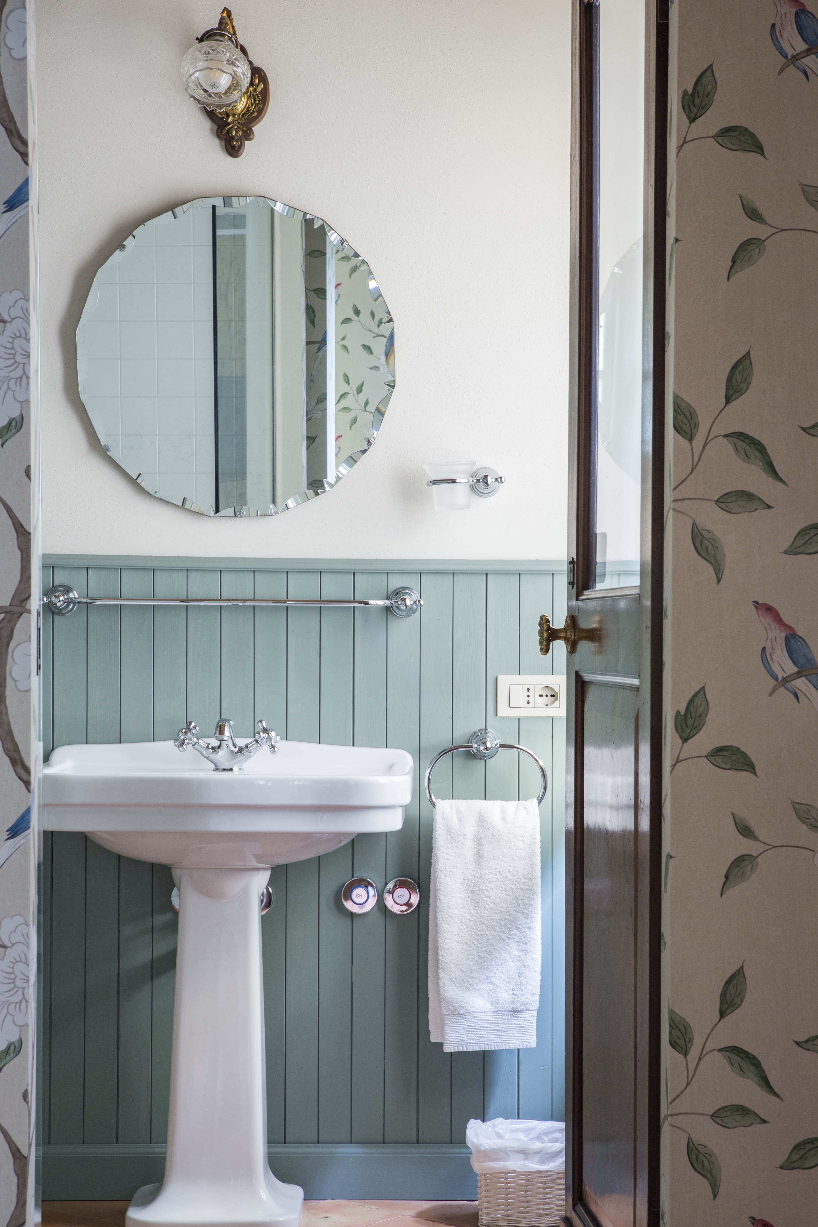 Elegant country-style bathroom with round mirror, pedestal sink and soft green paneling — boutique accommodation in rural Italy.