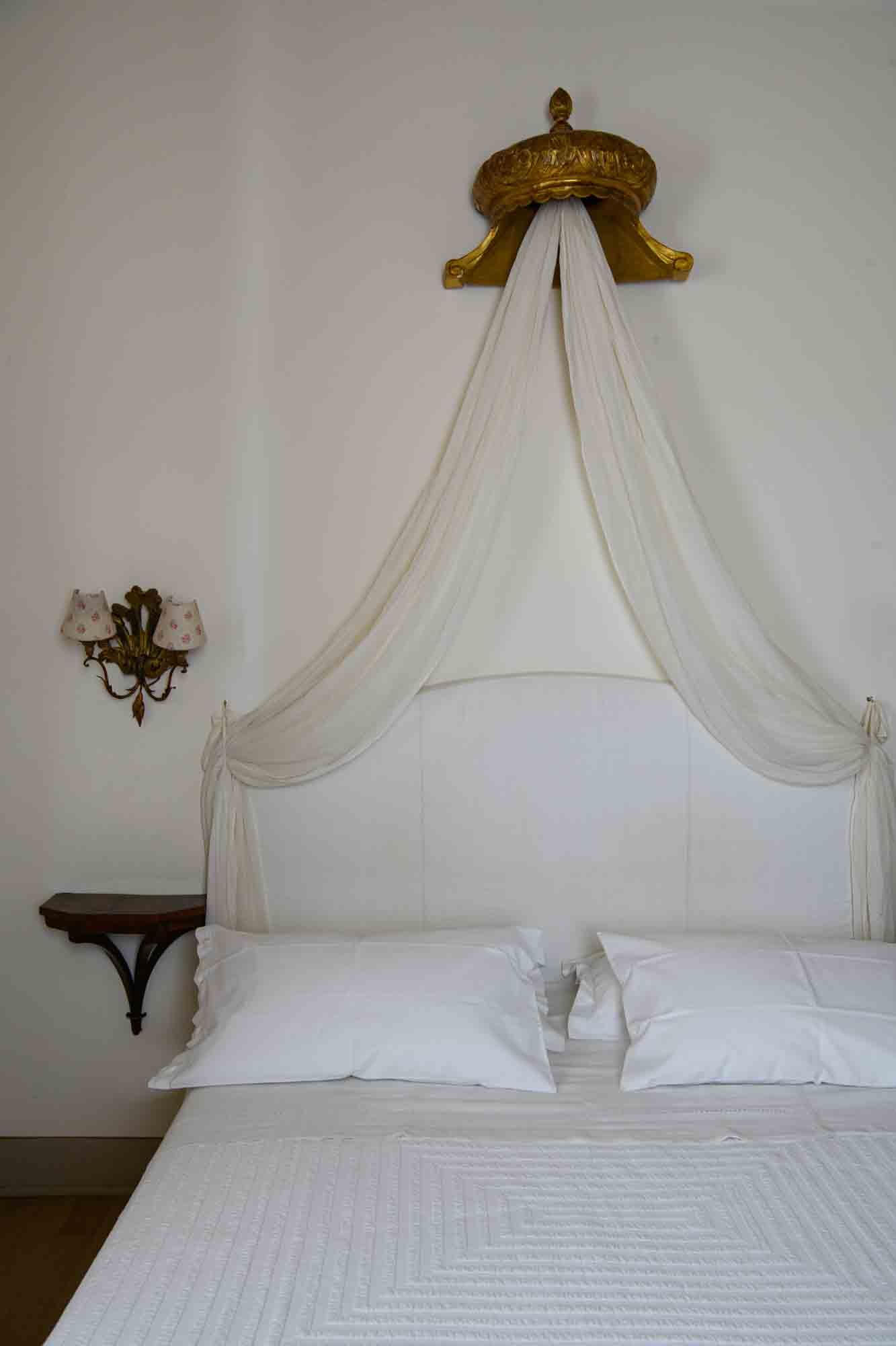 Elegant white bed with vintage canopy and antique wall lamps — serene Italian bedroom with timeless charm.
