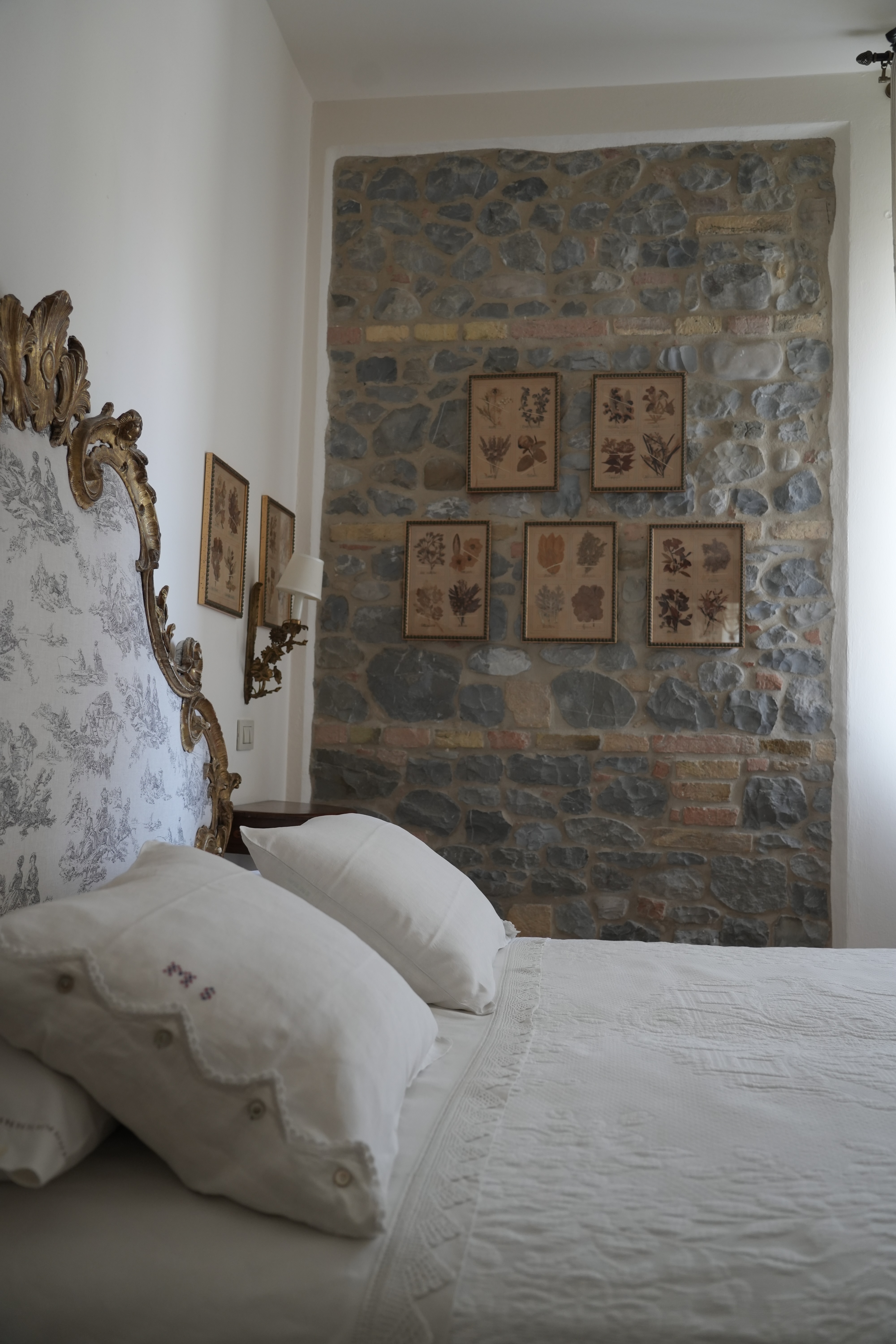 Elegant rustic bedroom with exposed stone wall, botanical prints and carved headboard — boutique accommodation in rural Italy.