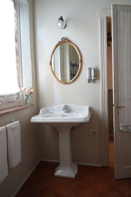 Bright vintage-style bathroom with pedestal sink, golden oval mirror and fresh flowers — charming rural accommodation.
