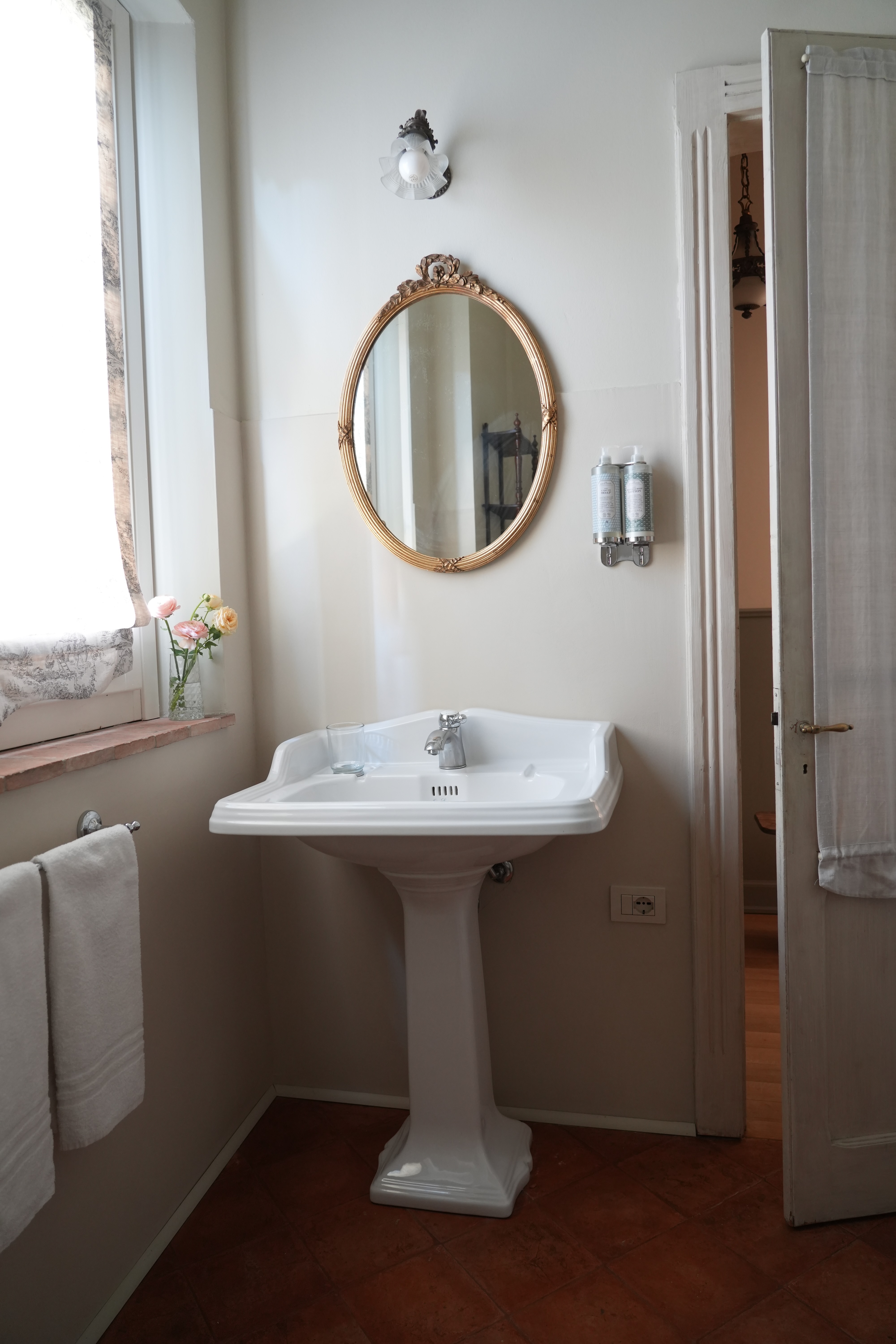 Bright vintage-style bathroom with pedestal sink, golden oval mirror and fresh flowers — charming rural accommodation.