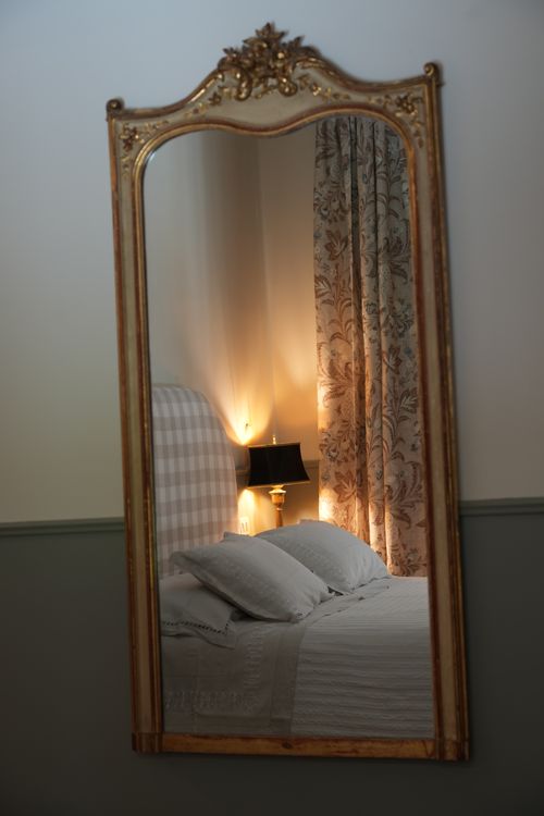 Reflection of a cozy bed with checkered headboard and floral curtains in an antique gold-framed mirror — Italian country chic interior.