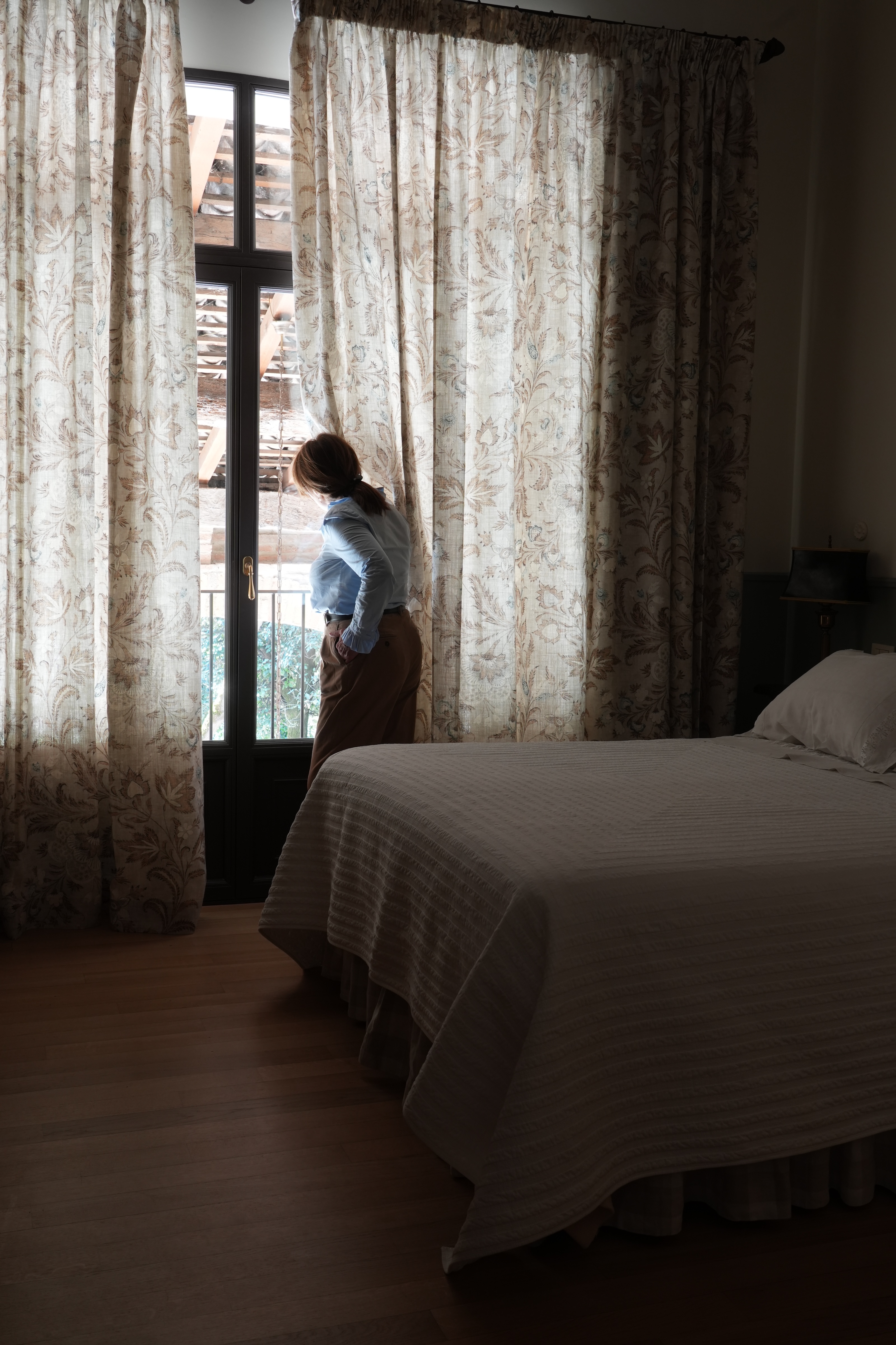 Woman looking out a window in a serene country bedroom with vintage floral curtains — peaceful boutique stay in Italy.