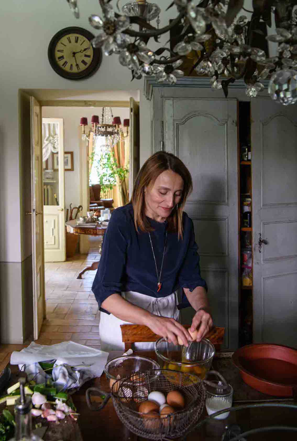 Woman cracking eggs into a glass bowl in a cozy kitchen with wooden furniture and a clock on the wall.