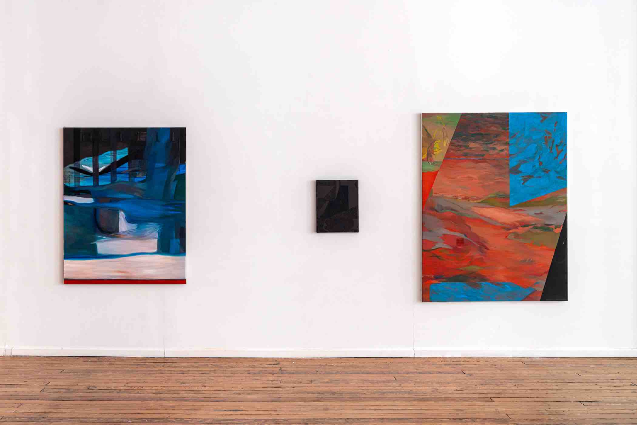 Three abstract paintings hanging on a white gallery wall above a wooden floor; left painting features blue and black tones, center painting is small and dark, right painting has red, blue, and orange hues.