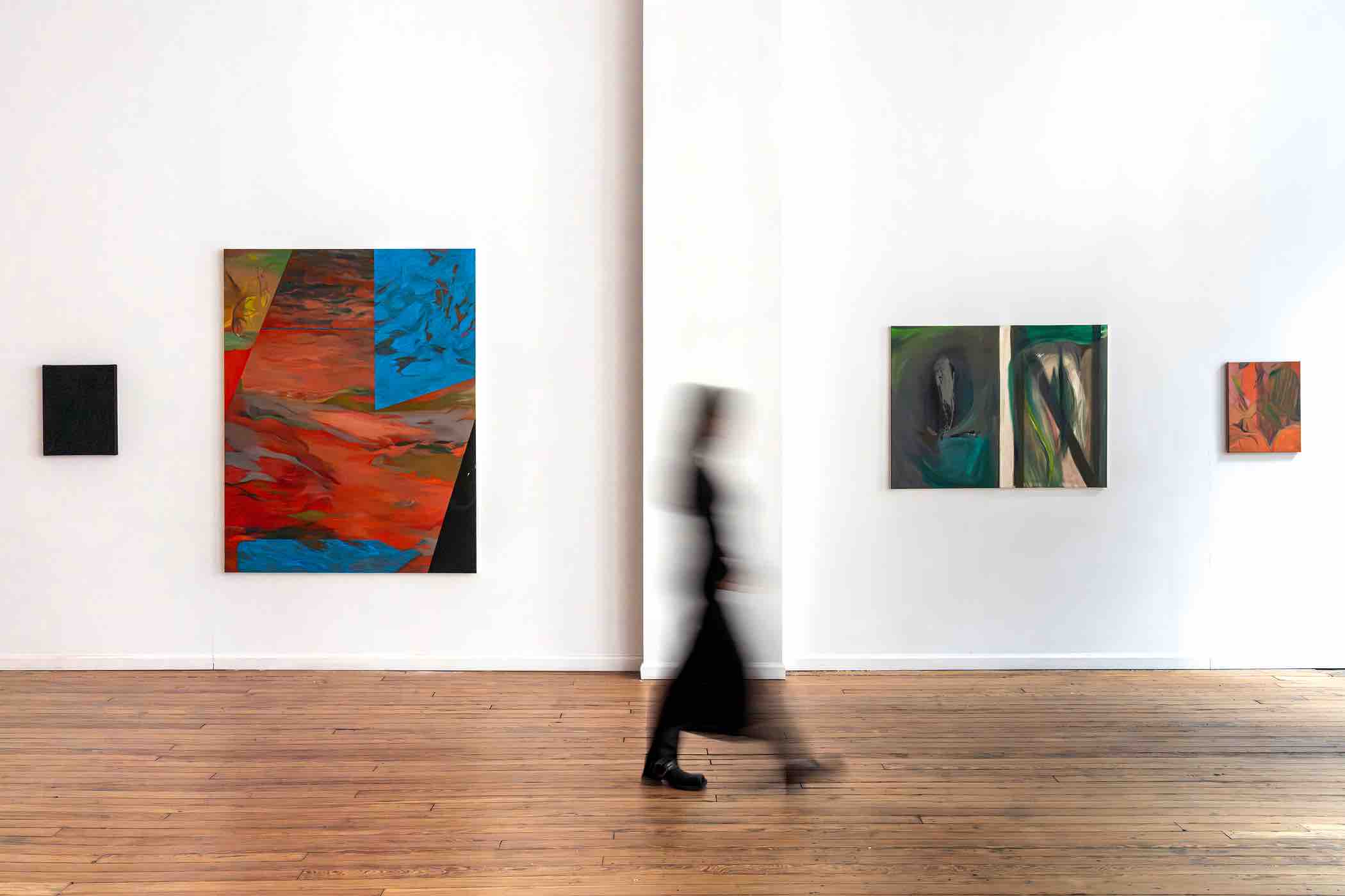 Blurred person in black walking through an art gallery with colorful abstract paintings on white walls and wooden floor.