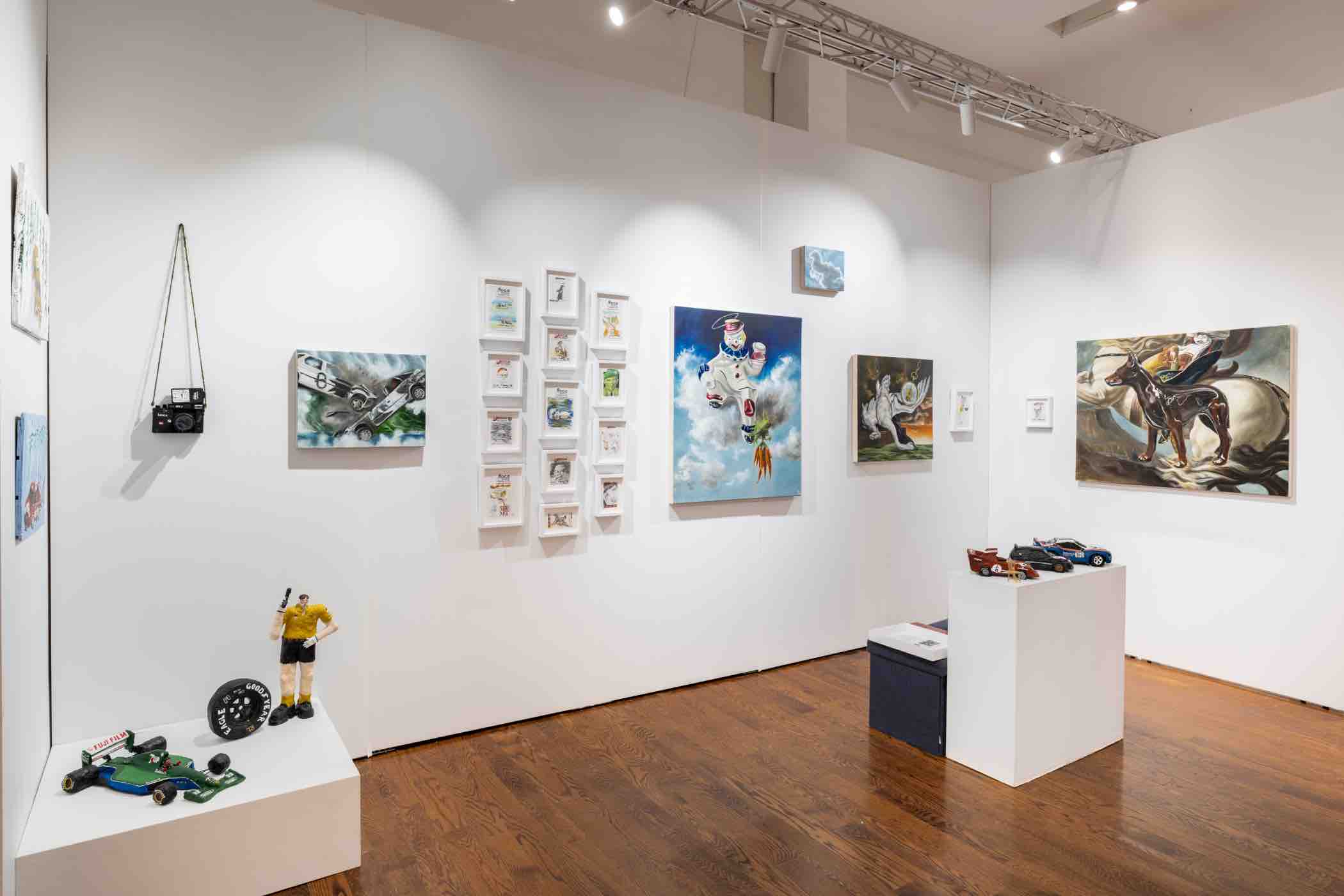 Gallery room with white walls displaying various colorful paintings and small sculptures on pedestals, including a toy race car, a figurine, and a model tire.