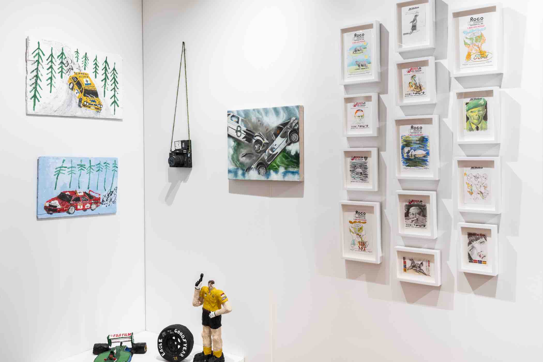 Art display with paintings of rally cars, framed sketches and paintings, a hanging black camera, and figurines including a man in a yellow shirt and a green model race car with a tire.