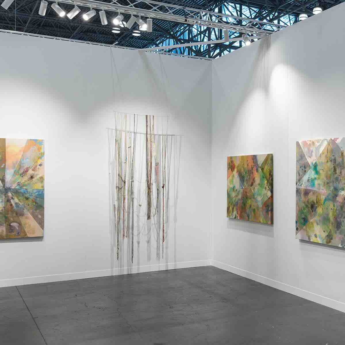 Corner of an art gallery with three abstract colorful paintings on white walls and an installation of thin, vertical hanging threads or strings illuminated by spotlights.