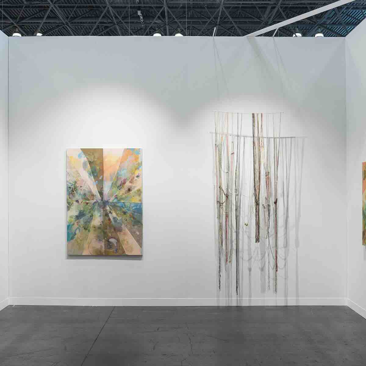 White gallery wall with abstract painting featuring radial patterns in muted pastel colors on the left, and a hanging art installation composed of long, thin vertical strands suspended in front of the wall on the right, casting shadows on the wall.