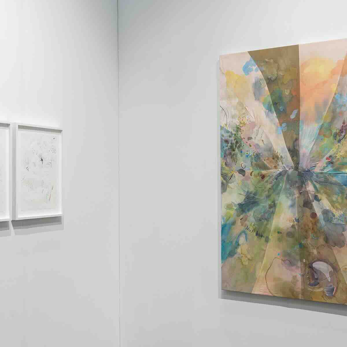 Abstract painting with a starburst pattern in soft pastel colors including greens, blues, yellows, and pinks on a white gallery wall, with two framed minimalist drawings on the adjacent wall.